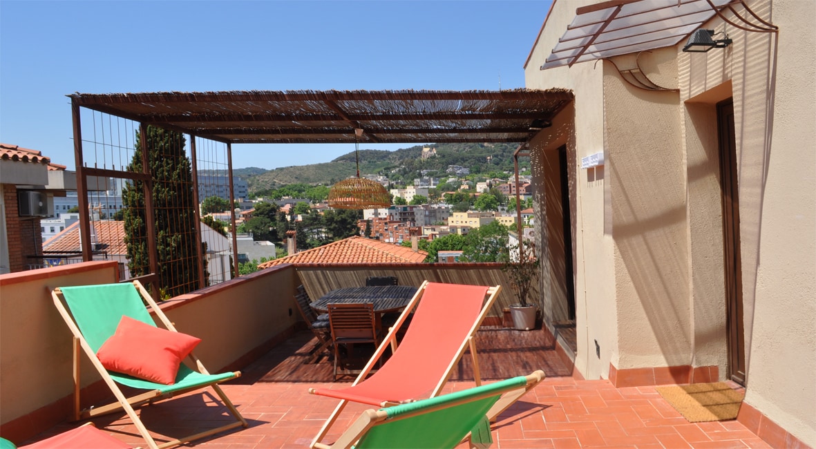 A spacious terrace is furnished with colorful lounge chairs and a wooden dining set. A pergola provides shade, enhancing the open-air experience. Scenic views of the surrounding greenery and rooftops are visible in the background, with bright blue skies overhead.