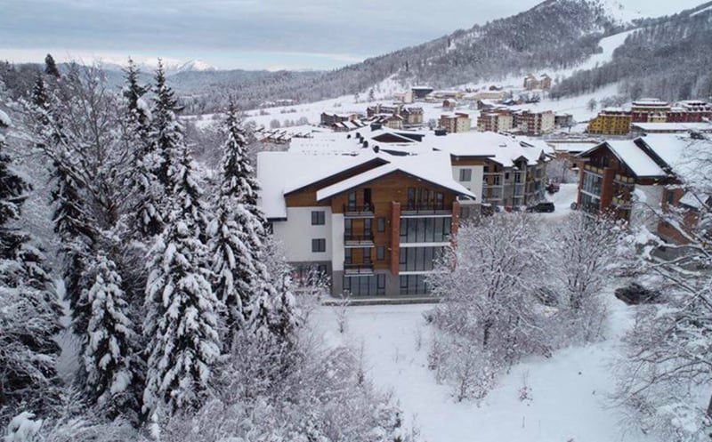 A snow-covered exterior of a modern apartment building is surrounded by tall coniferous trees. The landscape features a serene blanket of white snow, with distant mountains and additional buildings visible in the background, creating a peaceful winter setting.