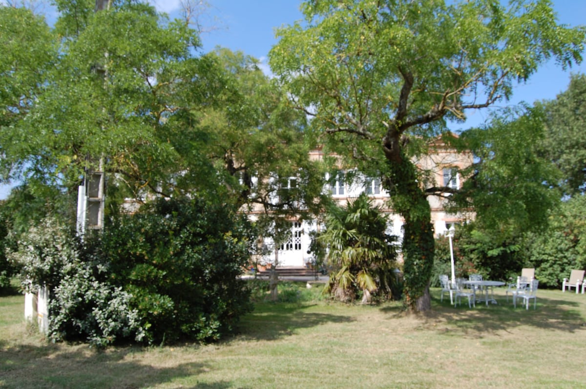 The exterior of a country house is surrounded by lush greenery, featuring flowering shrubs and large trees. A spacious lawn is visible, along with a patio area furnished with white chairs. The structure's large windows and rustic facade contribute to the tranquil countryside setting.