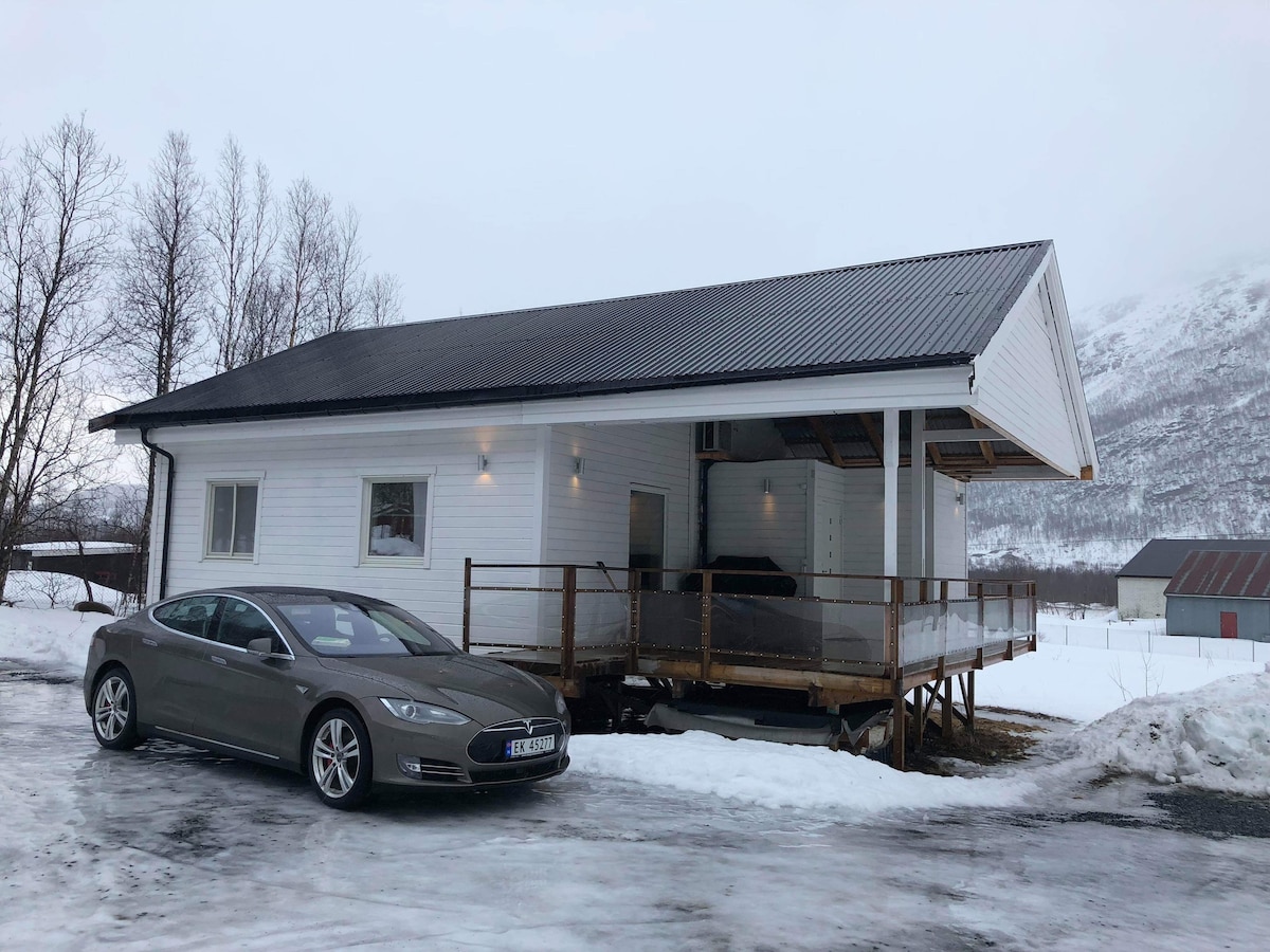 A modern white house is positioned next to a snowy landscape. The structure features a covered porch with large windows, blending indoor comfort with outdoor views. A silver car is parked in front, with mountains visible in the distance under a cloudy sky.
