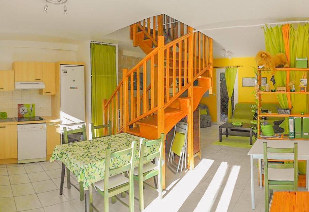 The open living area and kitchen feature a wooden staircase leading to the upper level. A dining table with green and beige accents is positioned beside the fully equipped kitchen. Colorful decor enhances the bright space, complemented by sunlight streaming through the large windows.