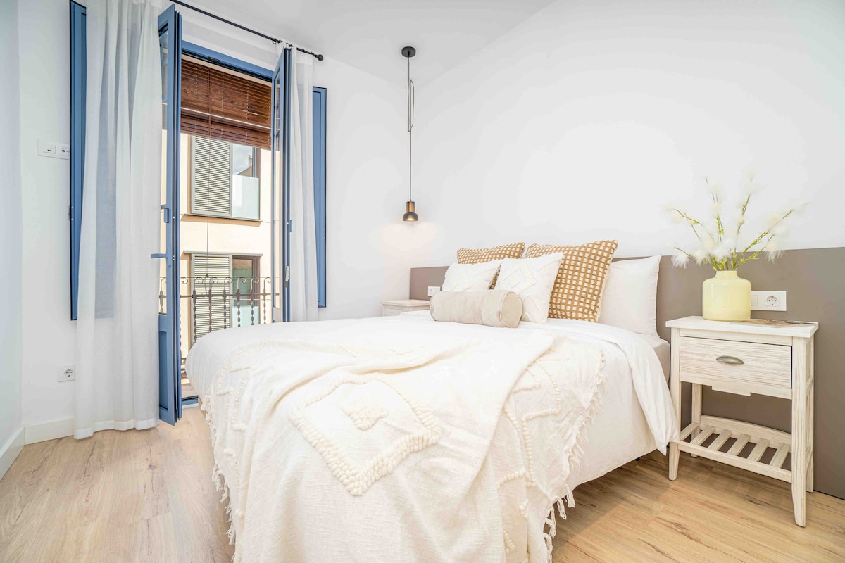 A bright bedroom features a spacious bed with neutral bedding and decorative cushions. A bedside table holds a small vase with greenery. French doors open to a balcony, allowing natural light to fill the space, complemented by light wood flooring.