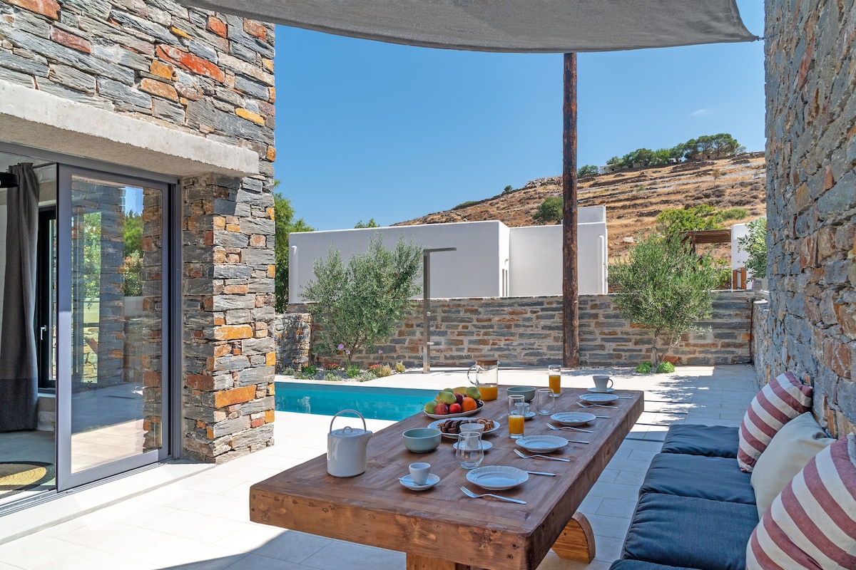 An outdoor terrace is presented with a wooden dining table set for breakfast. Plates, glasses, and a teapot are arranged alongside a bowl of fresh fruit. Olive trees are visible in the background, and a private pool glimmers under clear blue skies.