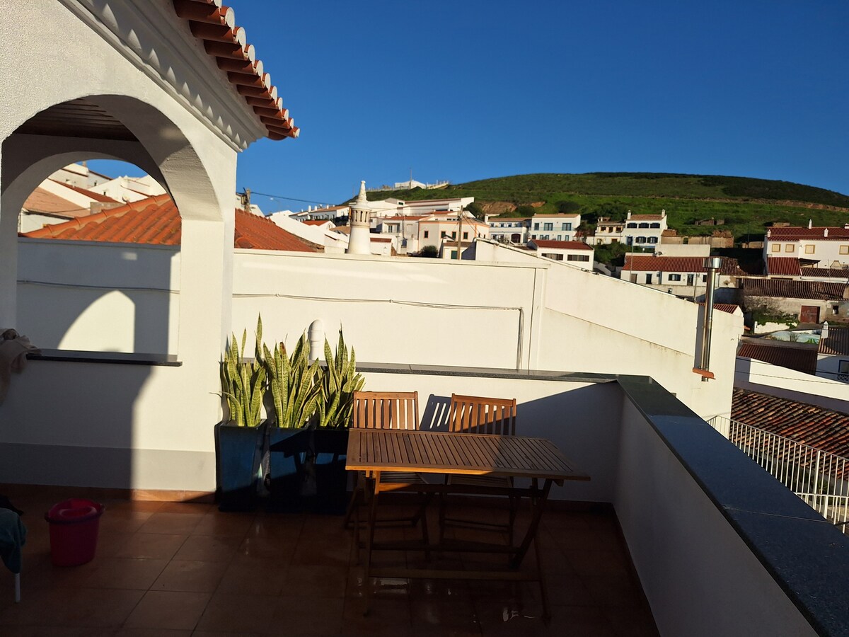 A private terrace is presented, featuring a wooden table and chairs positioned under a clear blue sky. Adjacent to the seating area, two large planters with greenery are visible. The backdrop showcases the village and hillside, creating a serene outdoor setting.