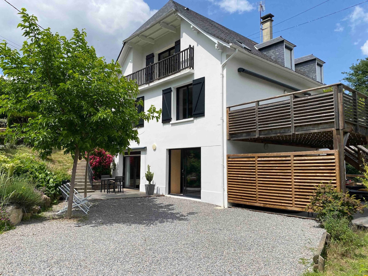 The exterior view of a three-story white house is highlighted, featuring a large balcony with wooden railings. A gravel area is visible in the foreground, accompanied by a dining set under a tree, while lush greenery surrounds the property.