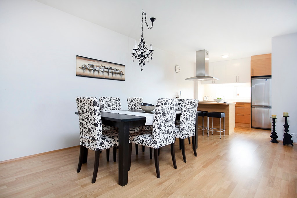 A dining area features a rectangular table surrounded by six patterned chairs. A chandelier hangs above the table, providing soft illumination. The adjoining kitchen is visible in the background, showcasing modern appliances and a clean design.