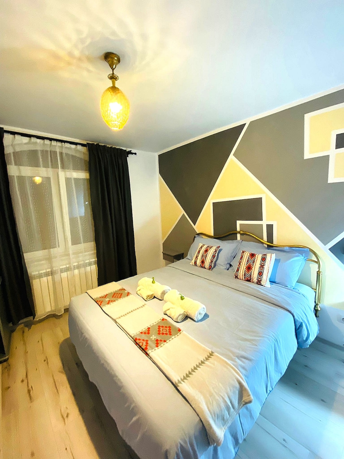 A cozy bedroom features a large bed with light linens and decorative pillows. A modern geometric mural adorns the wall behind the bed, while curtains frame the window allowing natural light to enter. A pendant light adds a subtle glow to the space.