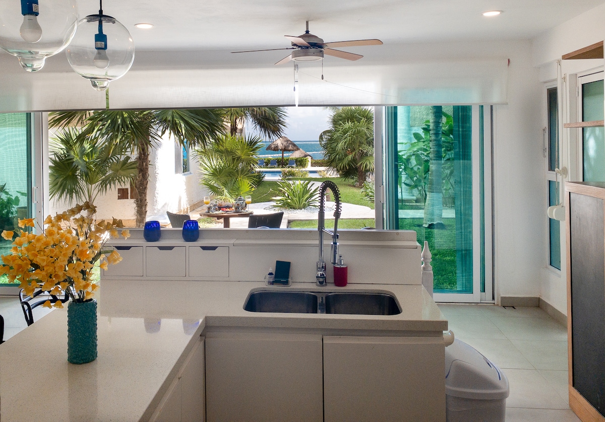 The kitchen features modern appliances and a large countertop, with a view of the garden and swimming pool through expansive glass doors. A vase of yellow flowers adds a bright touch to the space, while a ceiling fan enhances comfort.