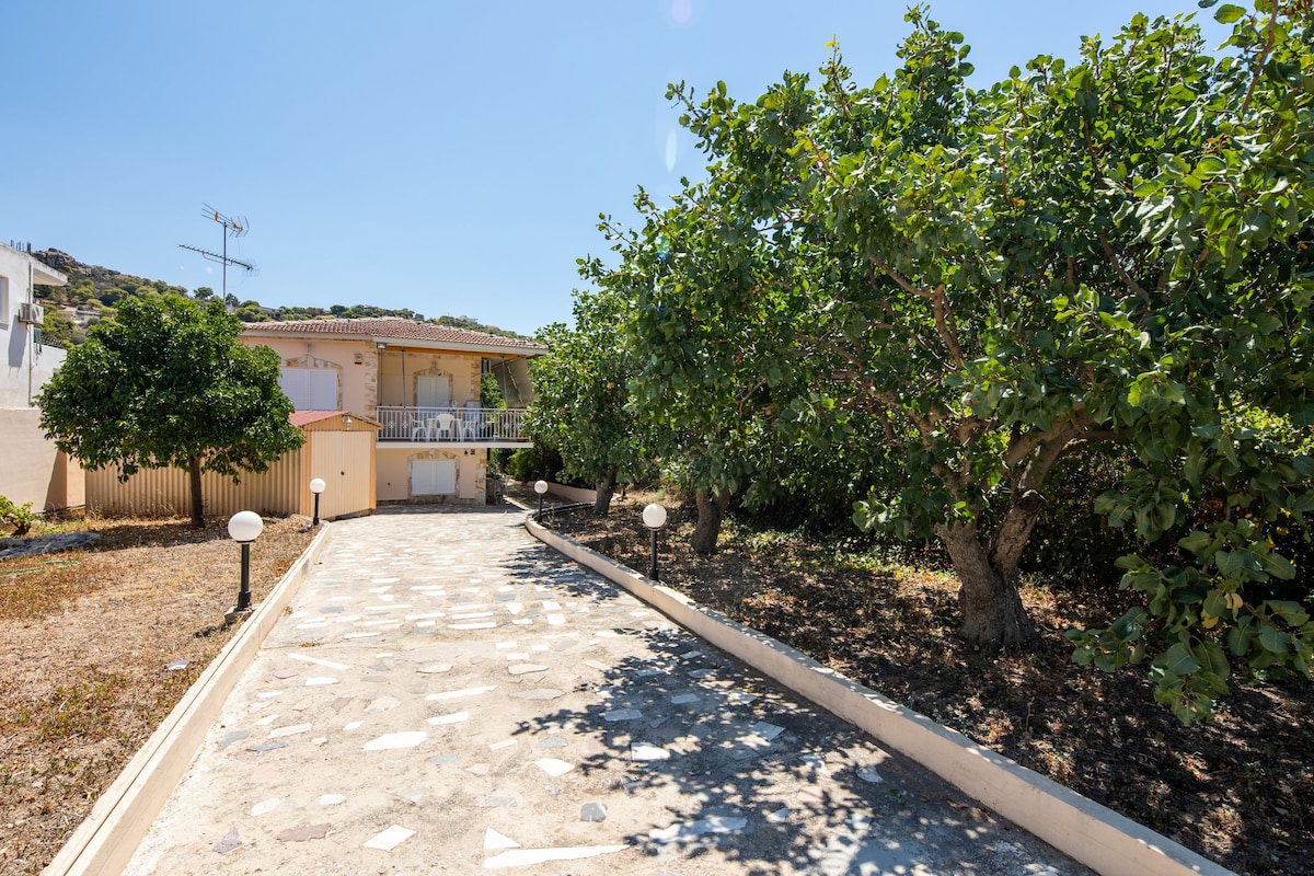 The entrance pathway leads to the apartment, framed by lush greenery and fruit trees. A spacious garden is visible on both sides of the stone path, and the building is slightly elevated with a balcony overlooking the area.