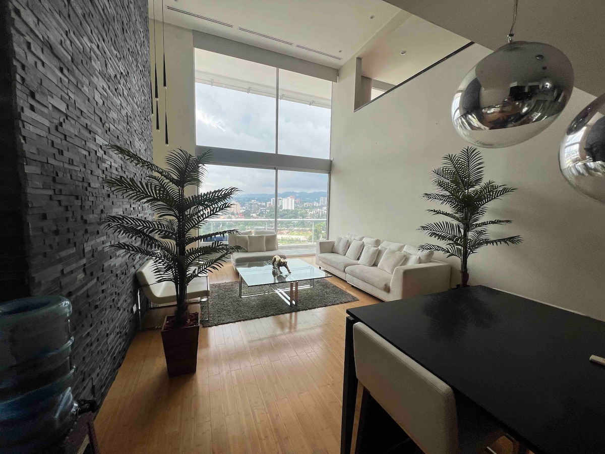 A spacious living area features large windows offering a panoramic view of the surrounding landscape. A comfortable white sofa is positioned around a glass coffee table, with tropical plants thoughtfully placed to enhance the decor. A dining table is visible, completing the open, airy feel.