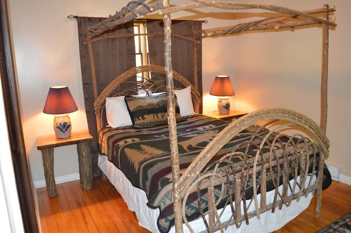 Chumba cha kulala cha kustarehesha cha nchi, kilicho na kitanda cha malkia cha mbao kilichoinama, standi ya usiku, kabati la nguo, na mlango wa kuteleza unaoangalia baraza la kujitegemea.