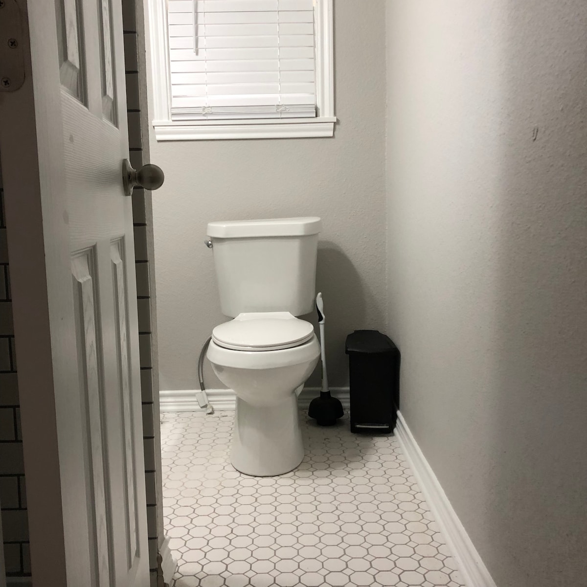 A clean and tidy bathroom space is seen, featuring a white toilet positioned against a light gray wall. A small black trash can and a toilet brush stand nearby, while natural light filters through a window with white blinds.
