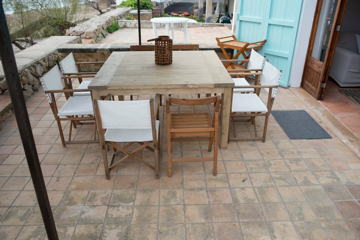 An outdoor dining area is presented with a wooden table surrounded by six chairs, crafted from light-coloured wood and white fabric. The serene patio is paved with square tiles, showcasing a natural setting with nearby greenery and a glimpse of the sea in the background.