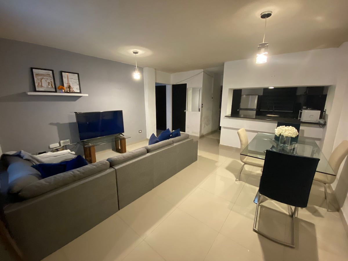 A modern living space is featured, showcasing a comfortable gray sofa adorned with blue cushions. A television is positioned on a wooden stand. Nearby, a dining area consists of a glass table accompanied by black chairs. The room exhibits neutral-colored walls and tiled flooring.