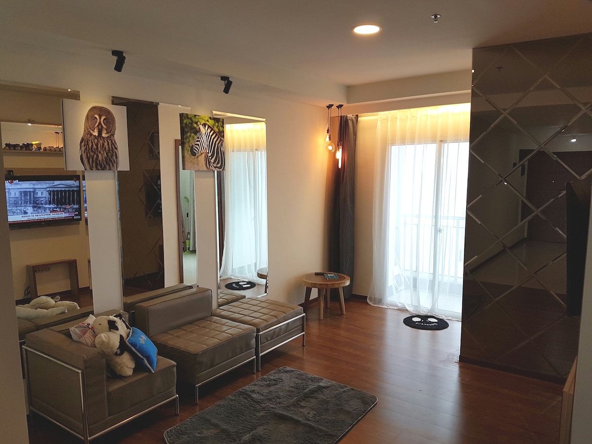A modern living area is displayed, featuring sleek furniture arranged around a small circular table. Natural light enters through large, floor-to-ceiling windows with sheer curtains. Decorative wall art adds character to the space, complemented by a plush rug on the wooden floor.