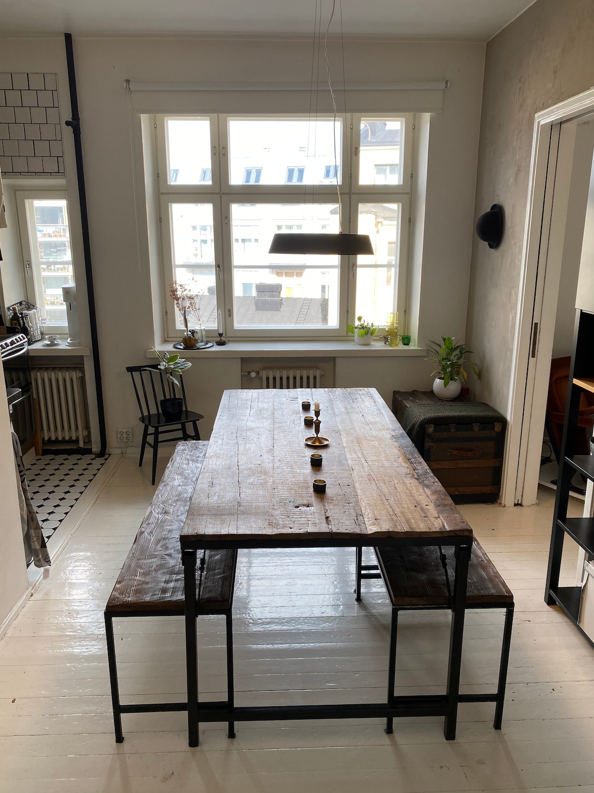 A spacious dining area features a large wooden table accompanied by two benches. Natural light floods the space through a set of wide windows. Simple decorations, including small plants and candles, add to the atmosphere. A modern pendant light hangs above the table.