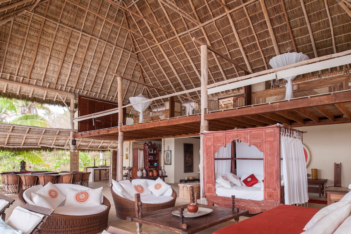 An expansive living area features a high palapa roof with natural straw, highlighting wooden beams. Cozy seating is arranged around ornate cushions, while a spacious outdoor bed draped in sheer fabric adds a touch of elegance. Lush greenery is visible through large windows.