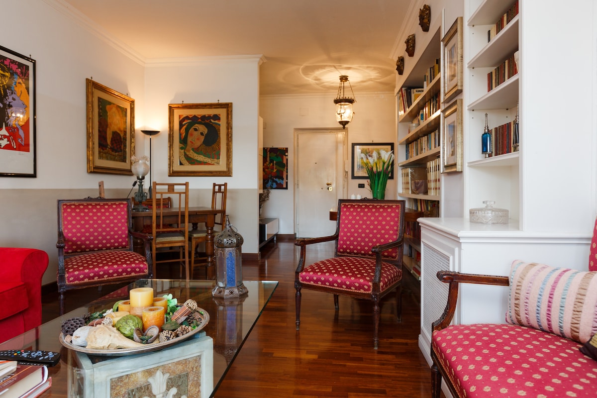 A comfortable living area is presented with two patterned armchairs and a red sofa. A coffee table, adorned with a decorative arrangement, stands at the center. The space features a wall of bookshelves and a dining area in the background, illuminated by soft lighting.