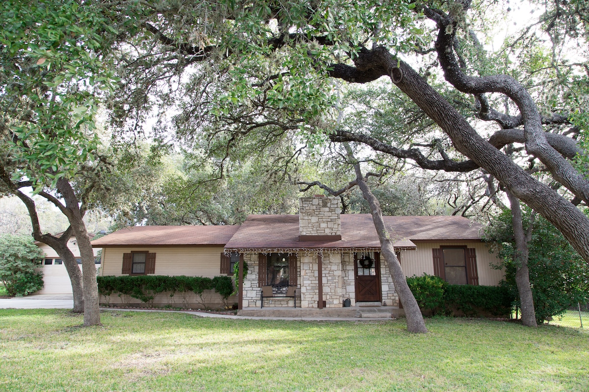 A charming single-story home is framed by mature oak trees, providing natural shade. A stone fireplace graces the front porch, which is accompanied by clear steps leading to the entrance. Lush green grass surrounds the property, enhancing the serene outdoor setting.