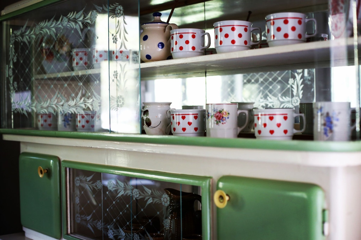 A charming kitchen display showcases an array of cups arranged neatly within a glass-fronted cabinet. The cups feature various designs, including red hearts and floral patterns. The cabinet's green accents complement the room's vintage aesthetic.