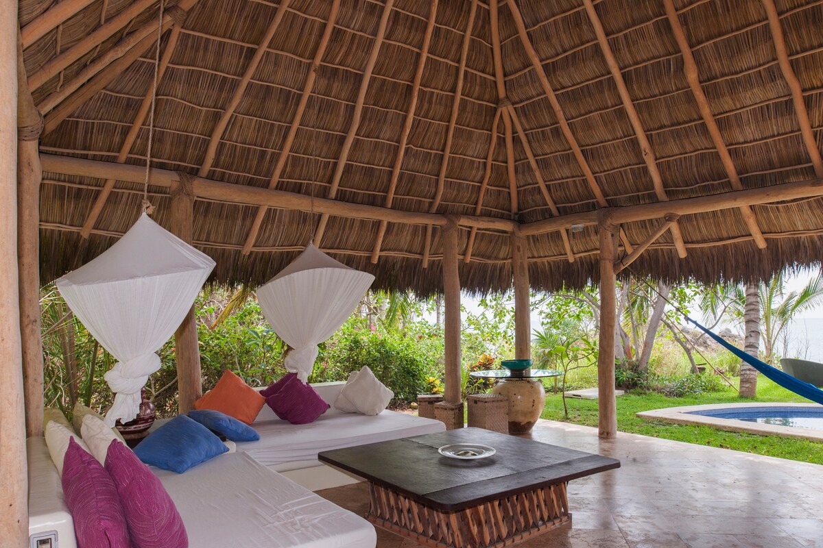 A shaded seating area features a thatched roof supported by wooden beams. Comfortable white couches with colorful pillows surround a central wooden table. A hammock can be seen in the background, alongside lush greenery and a glimpse of the ocean.