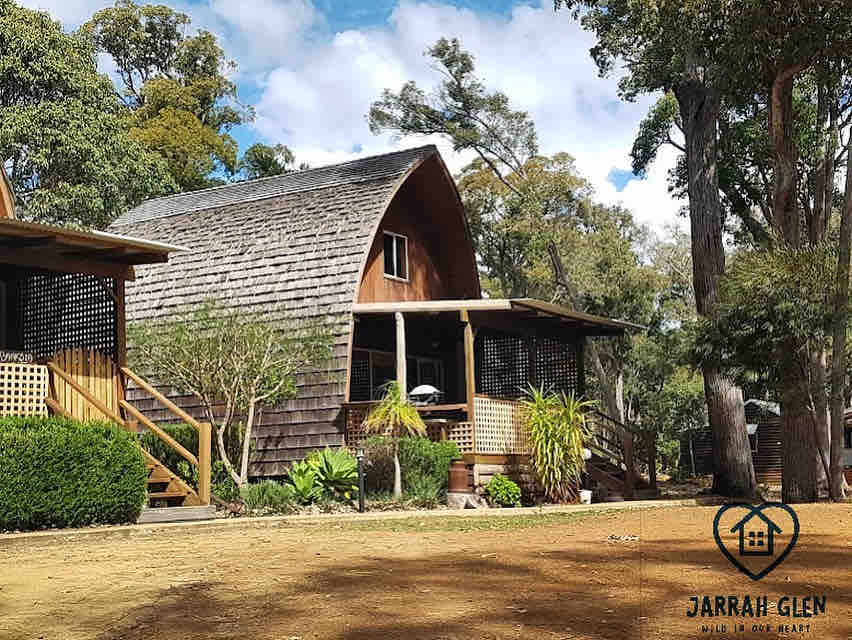 A wooden cabin with a curved roof is nestled among lush trees, showcasing a welcoming porch with railings and potted plants. Natural surroundings enhance the peaceful setting, inviting guests to relax and embrace the tranquility of the area.