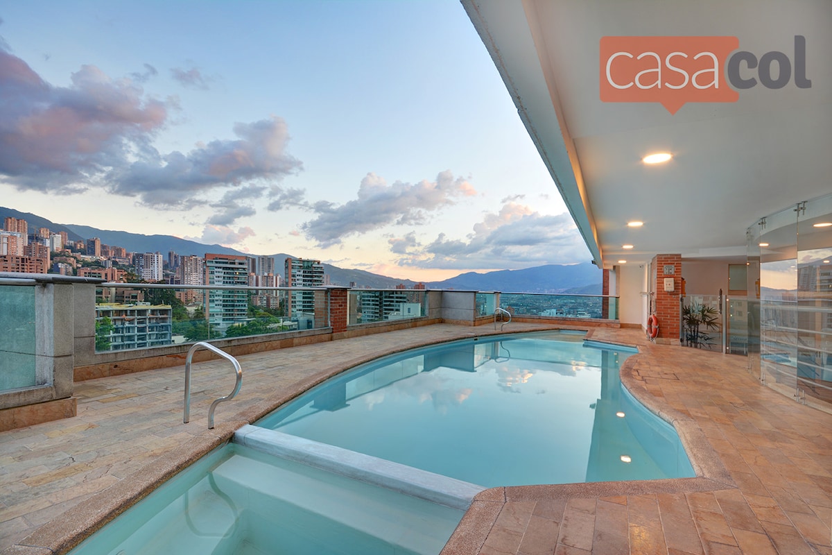 A rooftop pool area is depicted, featuring an inviting curved shape and clear water reflecting the sky. Surrounding mountains and cityscape are visible, complemented by a spacious deck area with smooth stone tiles. Sunset colors soften the scene, creating a calm ambiance.