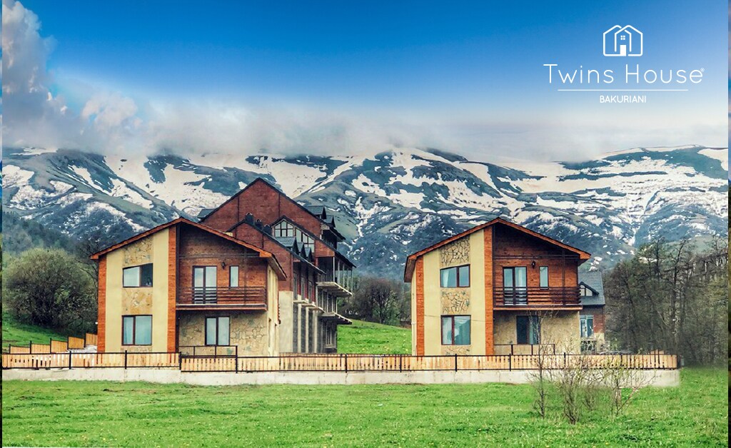 A charming two-building structure is set against a backdrop of snow-capped mountains. The buildings feature wooden and stone facades with balconies. Lush green grass surrounds the property, creating a serene outdoor setting. A wooden fence encloses the area, enhancing the rustic feel.