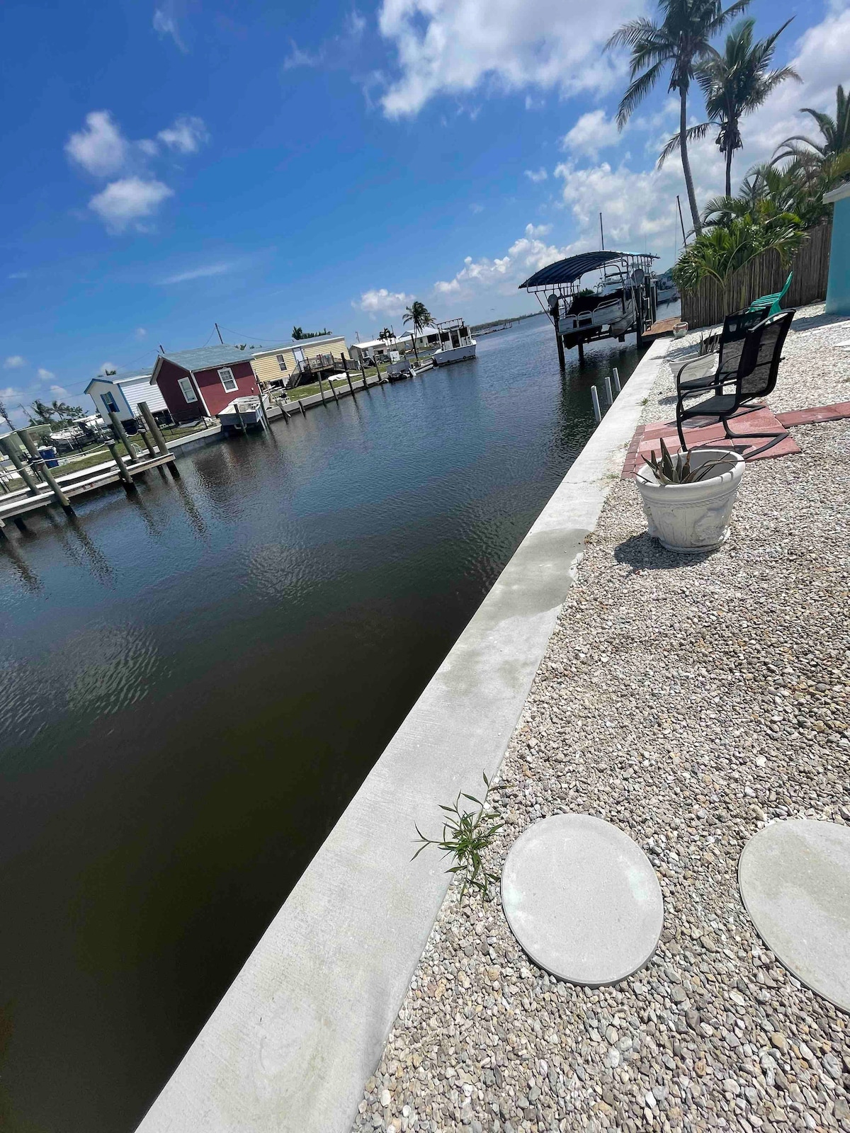 A tranquil waterfront scene featuring calm water reflecting a clear blue sky, with boats docked nearby. A concrete walkway runs alongside the water, bordered by pebbles and greenery. A black chair is positioned on the dock, offering a perfect spot for relaxation.