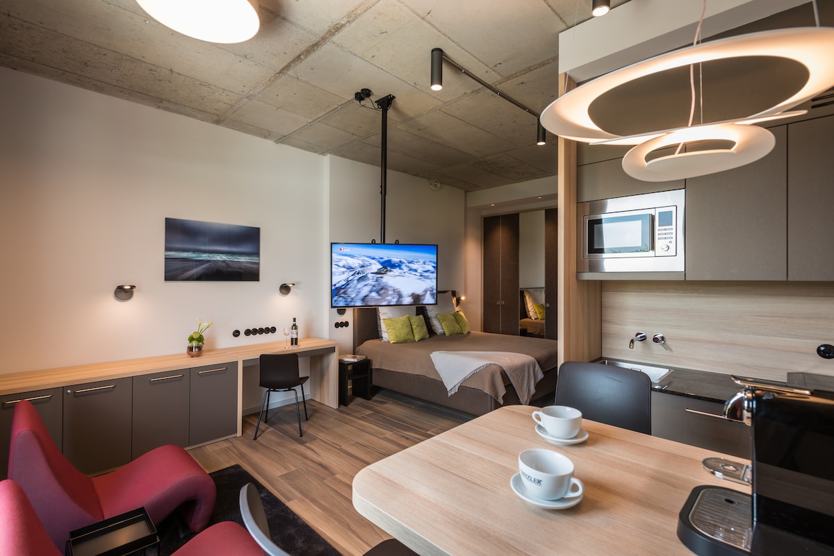 An open living area features a spacious bed with a neutral-colored blanket, complemented by a modern television mounted on the wall. A dining table with two cups is centrally located, and a fully equipped kitchen with a microwave is visible in the background.