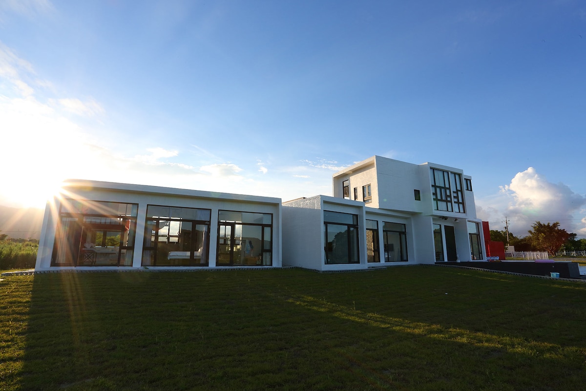 A modern, two-story building is surrounded by a lush, green lawn. Large glass windows dominate the facade, reflecting sunlight and offering views of the outdoor landscape. The design features clean lines and a minimalist aesthetic, enhancing the overall spaciousness of the structure.