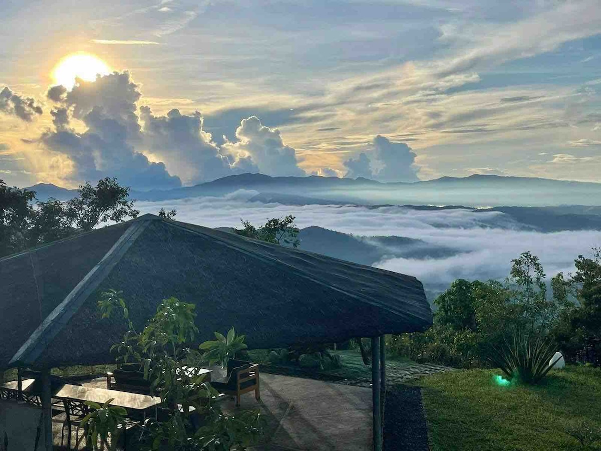 A gazebo is positioned against a backdrop of dramatic clouds and a rising sun, overlooking rolling hills. Below, a sea of clouds fills the valley, creating a serene atmosphere. Lush green foliage surrounds the area, providing a sense of tranquility and connection to nature.