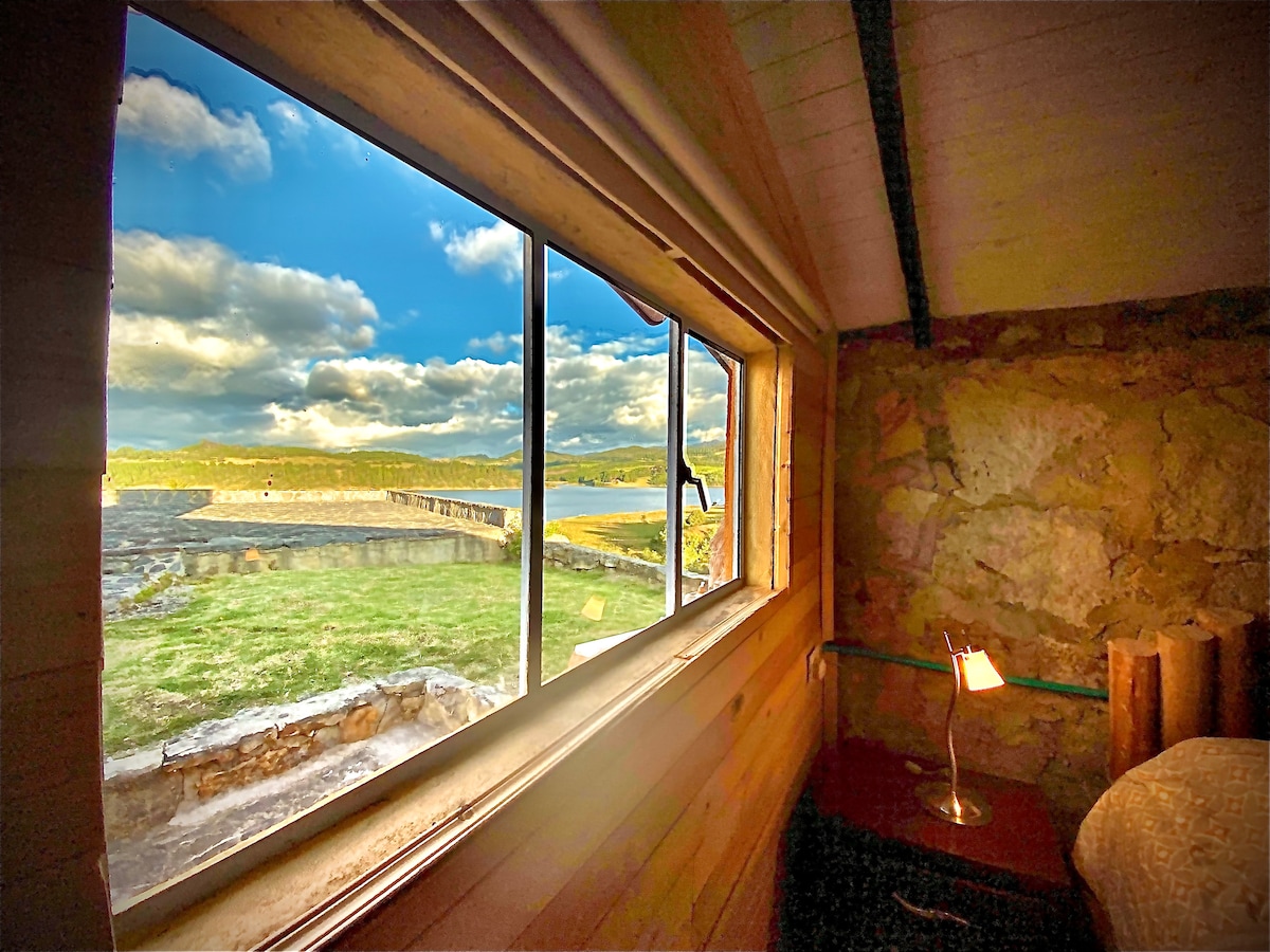 A window offers a view of lush green grass and the calm lake, with mountains visible in the distance. Natural light fills the room, reflecting off the stone walls. A bedside table with a lamp is positioned nearby, creating a cozy nook.