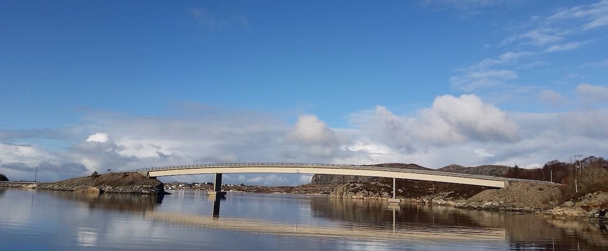 A modern bridge spans across calm waters, reflecting its arch in the clear blue surface. Surrounding hills are visible in the background under a partly cloudy sky, creating a serene landscape.