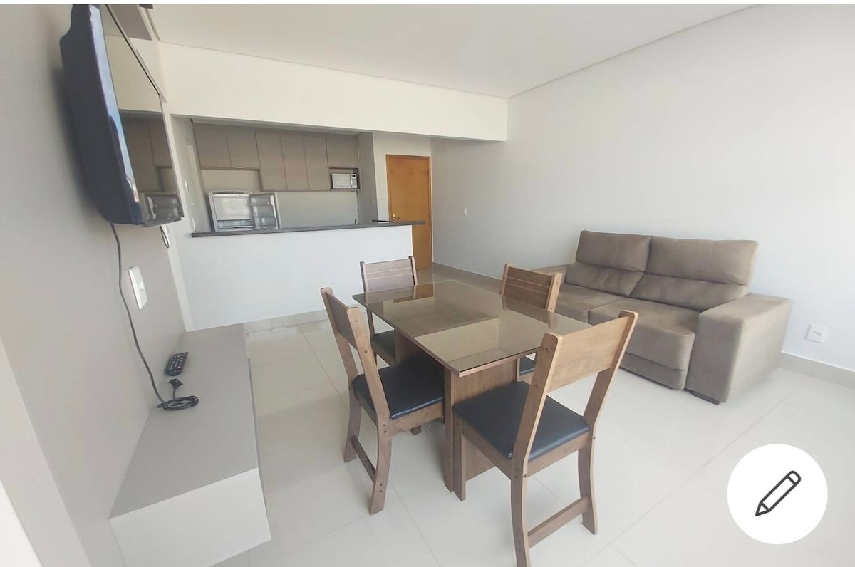 A modern dining area features a glass-top table surrounded by four wooden chairs, complemented by a comfortable sofa in the background. The open layout connects to a kitchen that is visible through a bar counter.