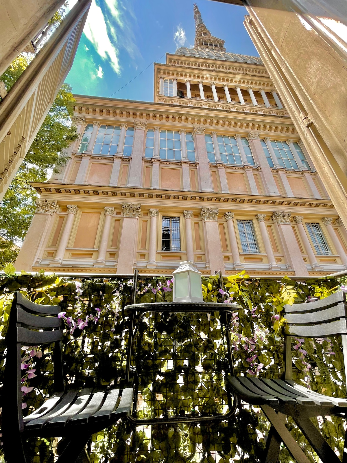 A charming balcony is viewed from the inside, featuring two black chairs and a small table. The space is adorned with greenery, creating a relaxed ambiance. Outside, the impressive Mole Antonelliana is visible, providing a picturesque backdrop.