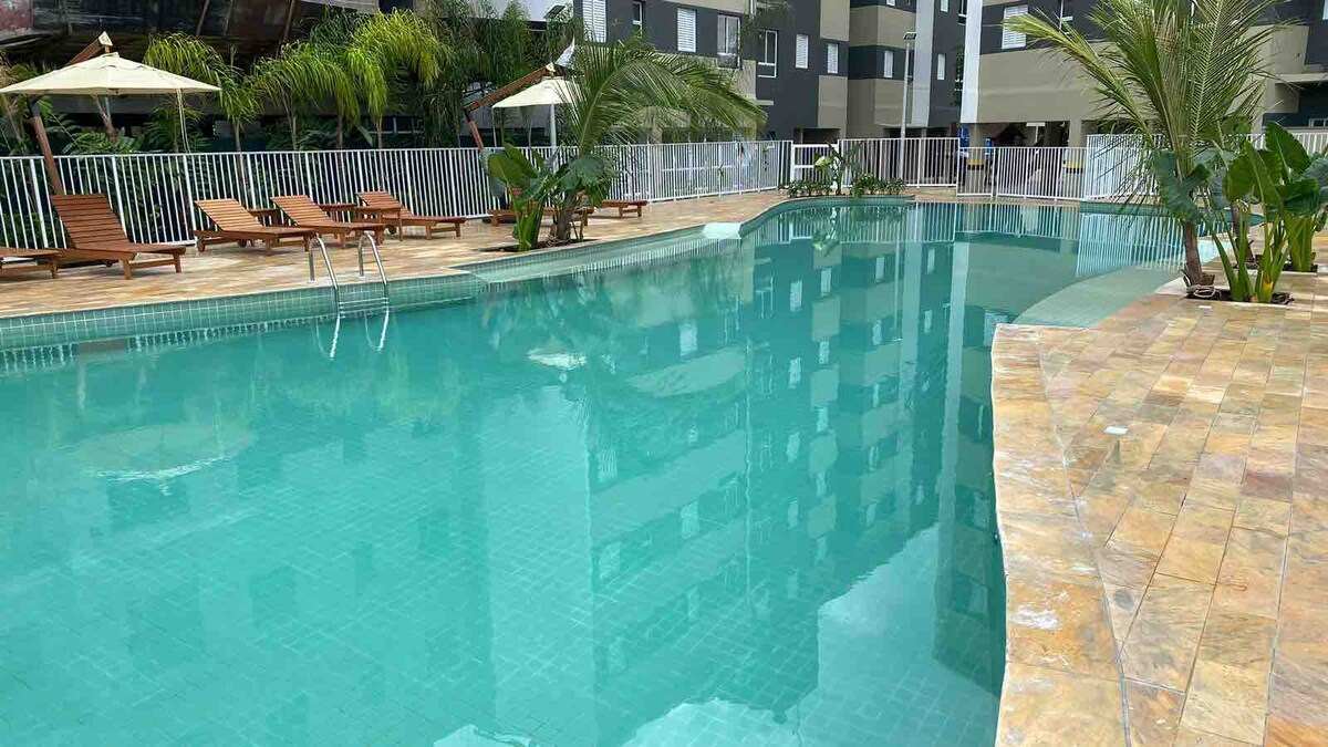 A spacious pool is surrounded by lounge chairs, with palm trees providing shade. Clear blue water reflects the surrounding buildings and greenery, creating a calm atmosphere for relaxation.
