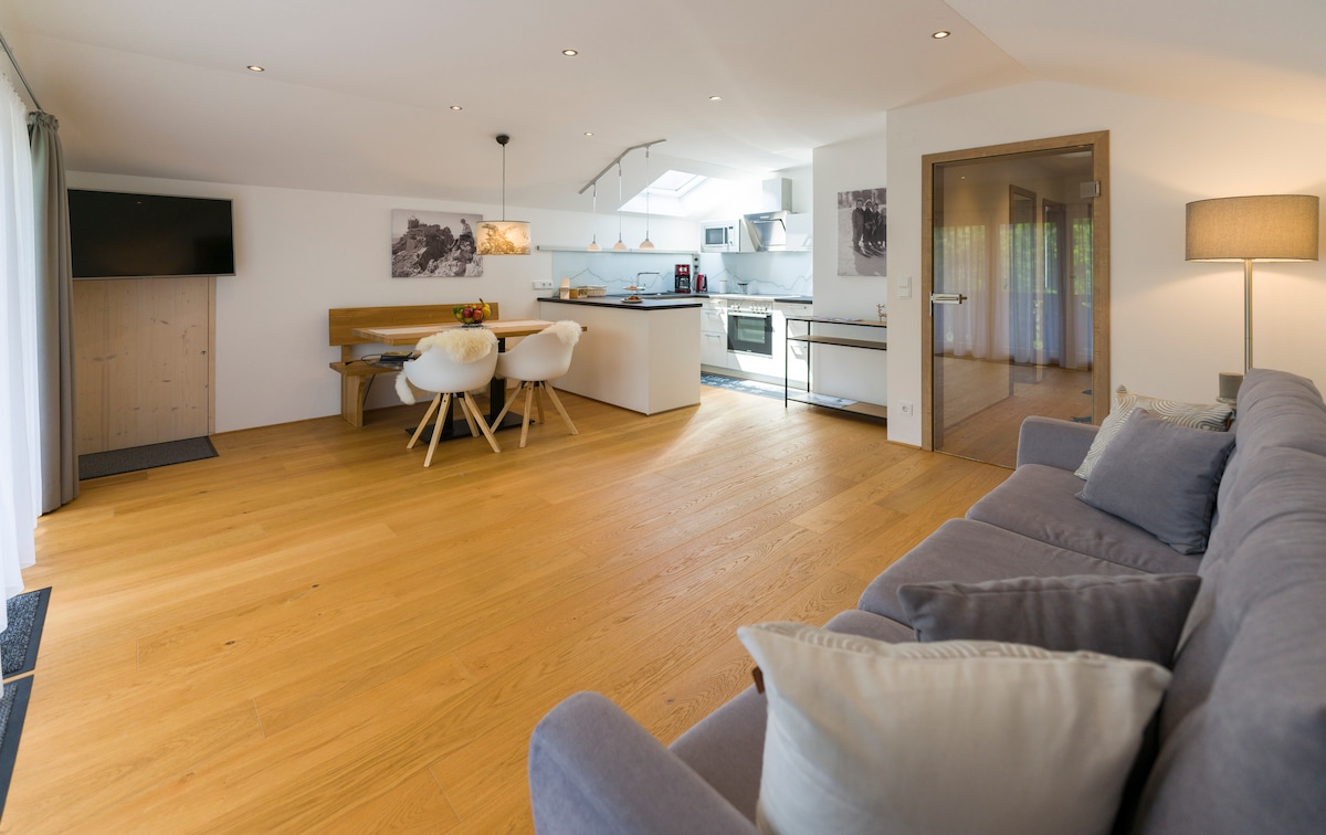 The open-plan living area showcases a light wooden floor with a spacious seating arrangement featuring a gray sofa. A dining table with white chairs and a fully equipped kitchen are visible, along with modern appliances and gentle lighting, creating a welcoming environment.