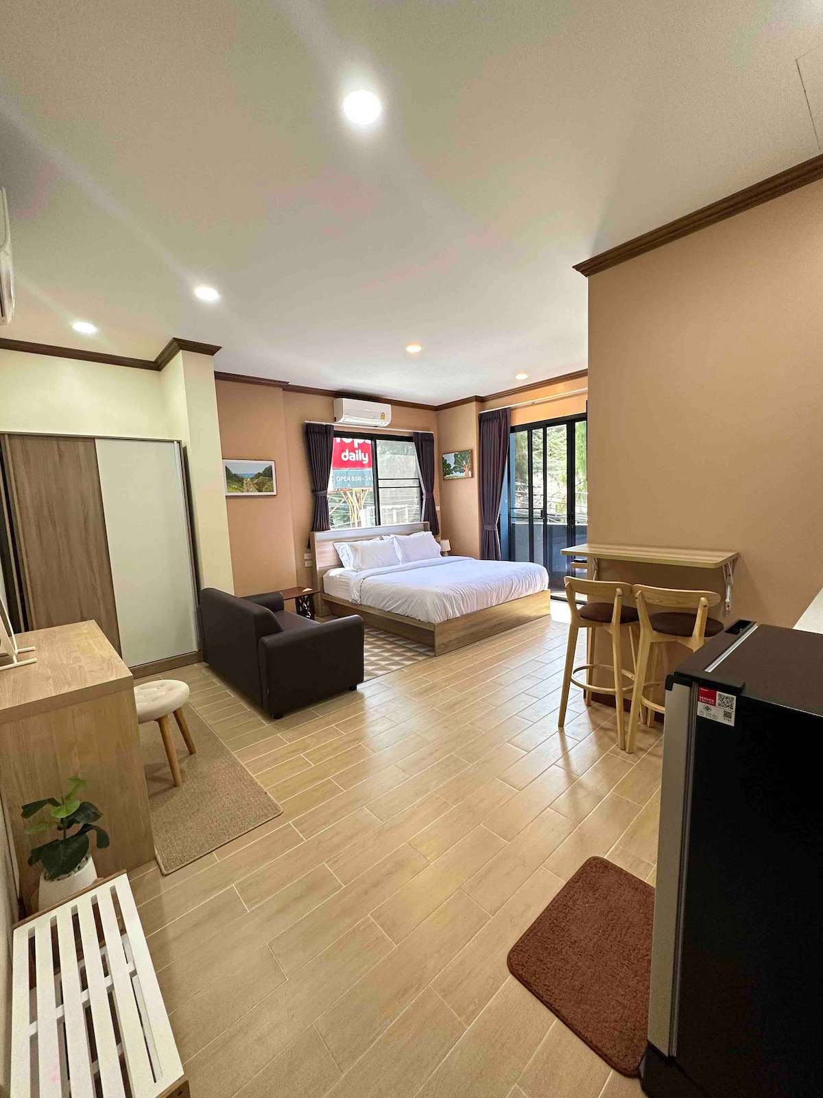 A bright room features a spacious bed, a compact kitchen area with a fridge, and barstool seating. Natural light is welcomed through large glass doors that open to a balcony. A sofa and a small dining table add to the room's functionality.