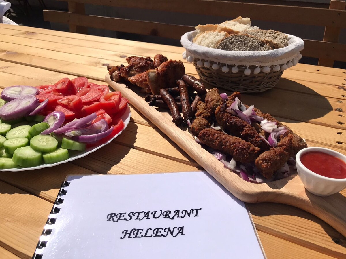 A wooden table is set with a variety of dishes, including grilled meats arranged on a wooden platter and fresh vegetables. A bowl of bread and spices is visible alongside a menu titled 'RESTURANT HELENA,' suggesting a casual dining experience.