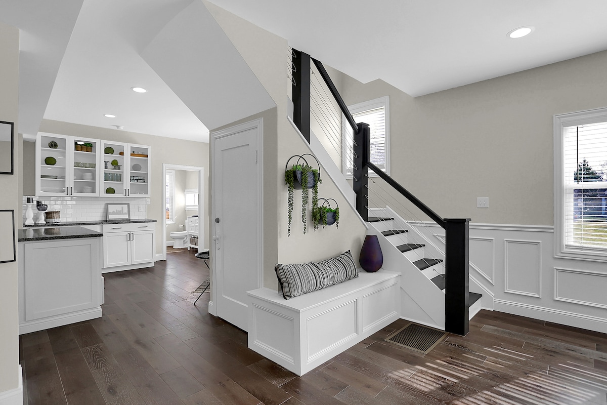A bright entryway features a stairway leading to the upper level, adorned with sleek black railings. To the left, a built-in bench offers seating beneath a hanging plant. The kitchen area is visible in the background, showcasing white cabinetry and modern appliances.