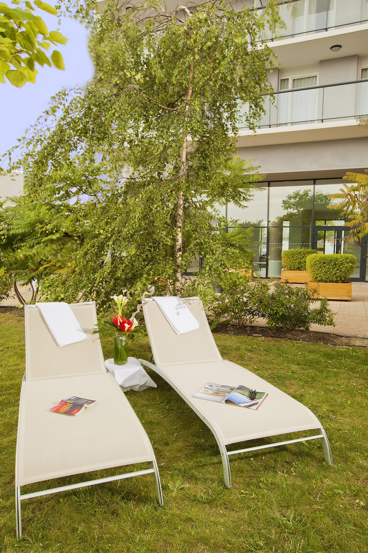 Two lounge chairs are positioned on a grassy area, accompanied by a small table with a flower arrangement. A tree provides shade, while the modern building is visible in the background, featuring large glass windows that allow for an open view of the outdoors.