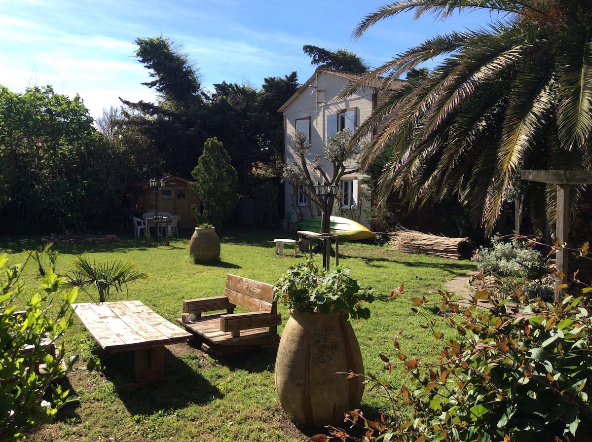 A spacious garden is featured, showcasing greenery and a mix of plants, including palm trees. Wooden seating is arranged around a rustic table, inviting relaxation. The two-story chalet in the background is surrounded by nature, providing a peaceful outdoor atmosphere.