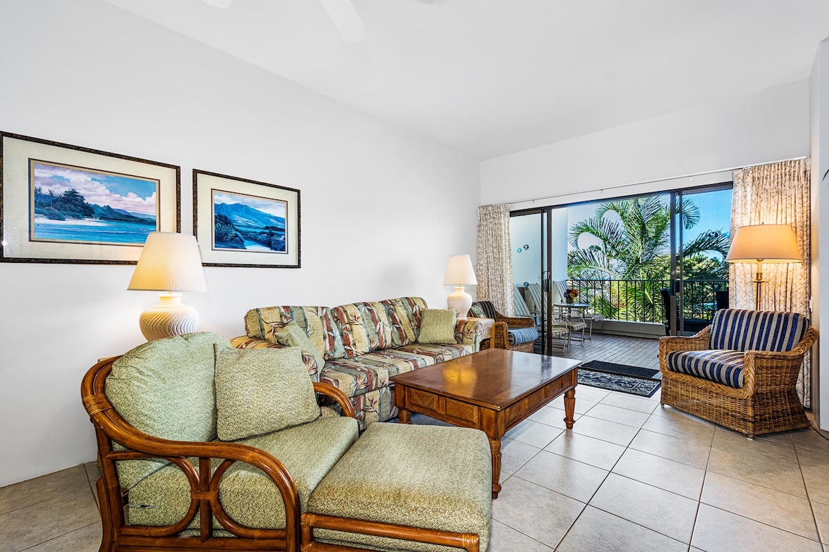A bright living area is presented featuring a light-colored tile floor and large glass doors offering views of lush greenery. A comfortable sofa with tropical patterns is complemented by two armchairs and a wooden coffee table. Soft lighting is provided by two table lamps.