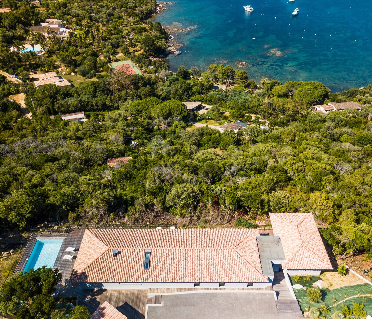 An aerial view captures the villa nestled amidst lush greenery with the turquoise waters of the Gulf of Ajaccio in the background. A private swimming pool is visible beside the villa, and boats can be seen in the distant sea, enhancing the coastal scenery.