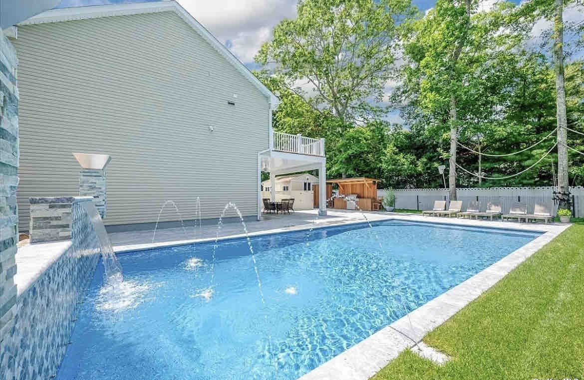 An in-ground pool is showcased, featuring decorative water features that create gentle splashes. Lush green grass surrounds the pool area, with lounge chairs positioned nearby. A shaded deck is visible in the background, along with a wooden structure for additional seating.
