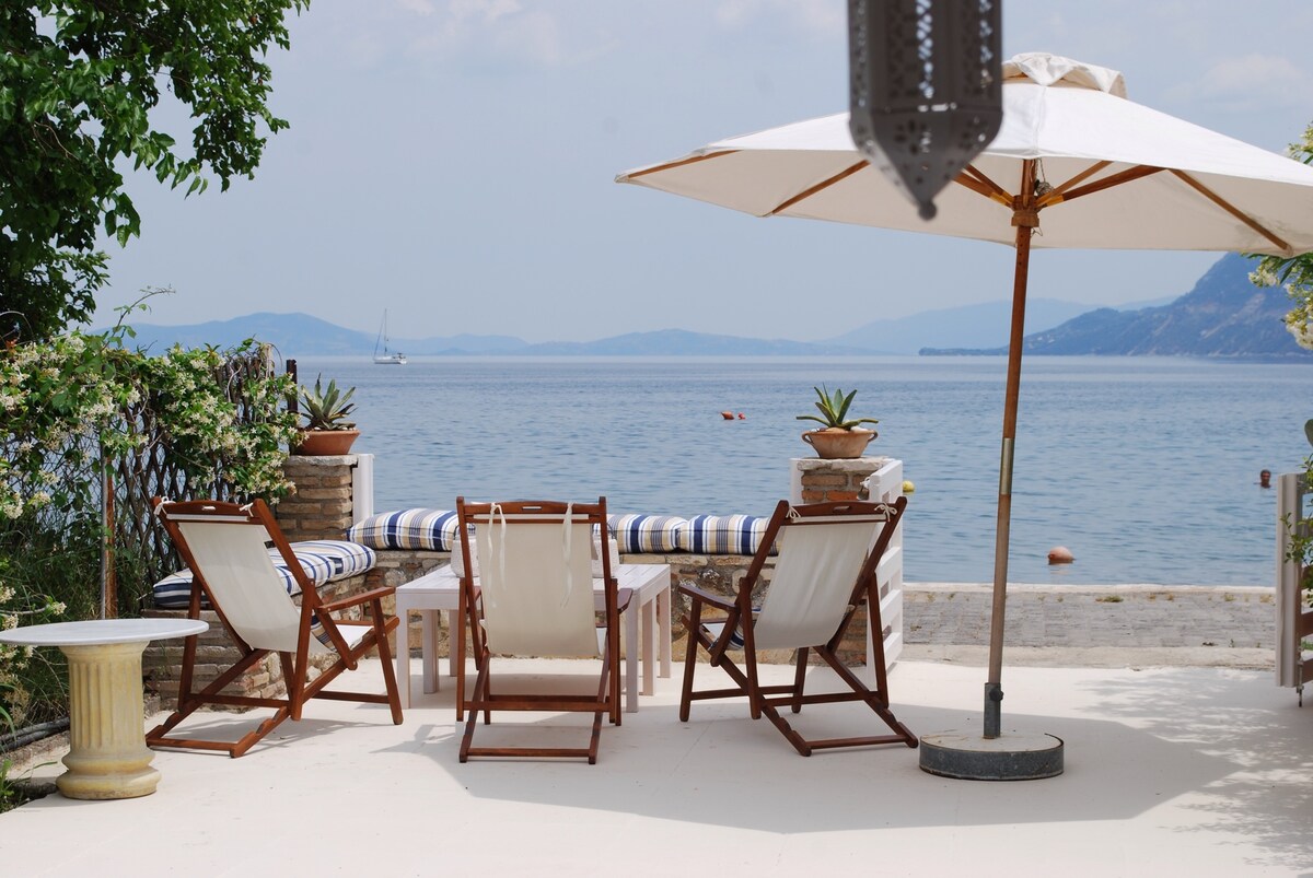 A serene outdoor seating area features two wooden lounge chairs and a small table, shaded by a large umbrella. Potted plants add greenery, while a view of the sea and distant hills creates a tranquil backdrop.