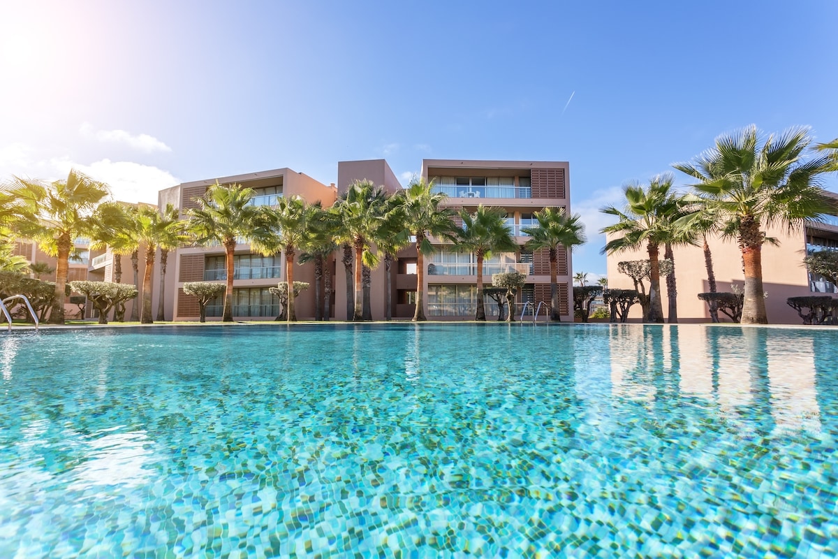 A large, inviting swimming pool reflects the clear blue sky, surrounded by well-maintained palm trees. The modern apartment buildings in the background feature expansive glass balconies, providing a view of the tranquil pool area. Sun loungers are visible along the pool's edge.