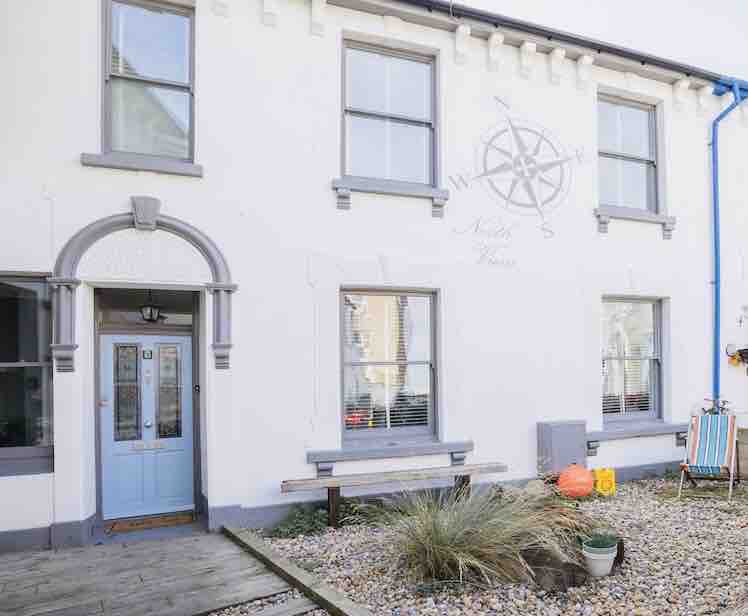 The exterior of the holiday home features a charming façade with a light blue door, framed by symmetrical windows. A decorative compass motif is displayed on the wall, while a stone path leads to a low bench area surrounded by gravel and coastal plants.