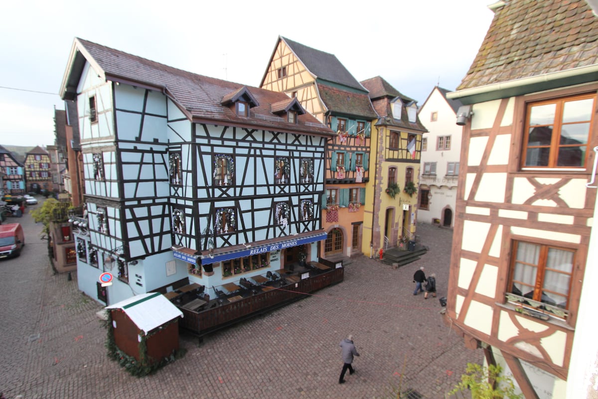 A charming square is surrounded by traditional half-timbered buildings. The pastel-colored facades and detailed wooden structures add character to the street. Cobblestone pathways lead through the area, with a few pedestrians walking leisurely. Outdoor seating is visible at the base of one building.